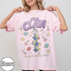 Puede incluir: Una camiseta rosa claro de manga corta con un estampado gr&aacute;fico de tres alien&iacute;genas verdes colgando de una garra. El texto dice THE CLAW y THE MYSTIC PORTAL AWAITS, rodeado de ilustraciones de cohetes, planetas y estrellas de colores.