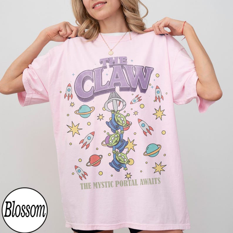 Puede incluir: Una camiseta rosa claro de manga corta con un estampado gr&aacute;fico de tres alien&iacute;genas verdes colgando de una garra. El texto dice THE CLAW y THE MYSTIC PORTAL AWAITS, rodeado de ilustraciones de cohetes, planetas y estrellas de colores.