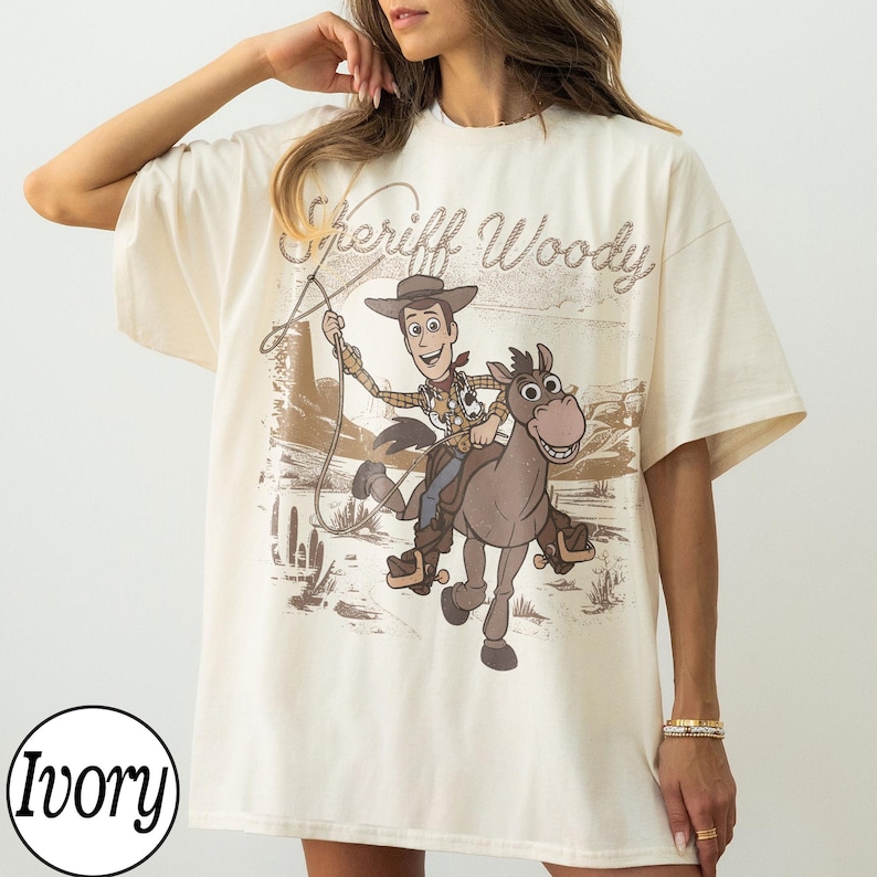 May include: Ivory-colored t-shirt featuring a graphic of Sheriff Woody riding a horse, with the text "Sheriff Woody" above the image. The design is in shades of brown and tan, with a desert landscape backdrop.