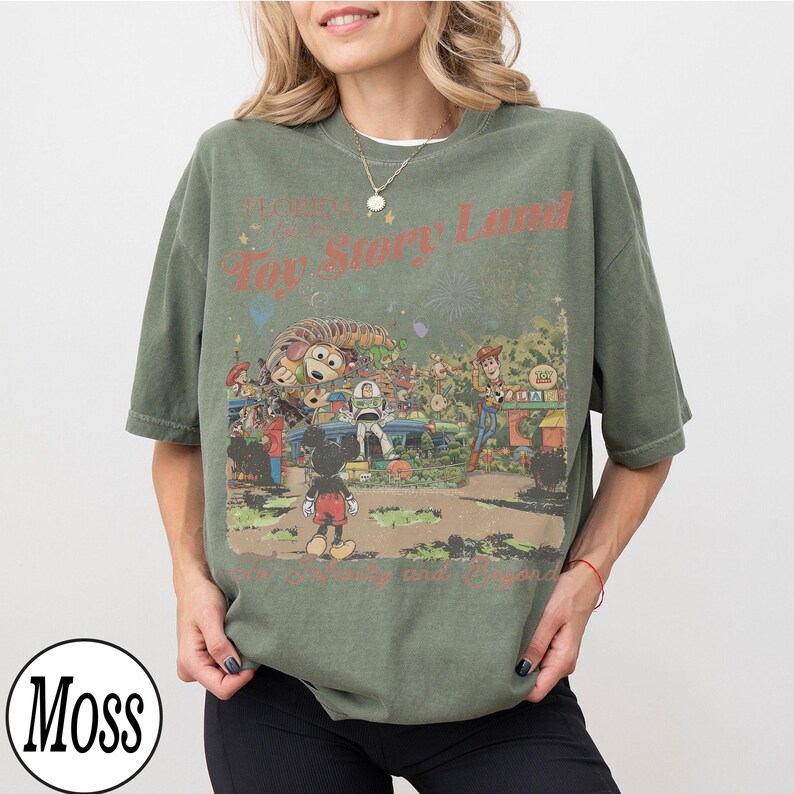 May include: An olive green t-shirt with a retro Toy Story Land graphic. The design features Mickey Mouse and other characters, with the words "Toy Story Land" in red.
