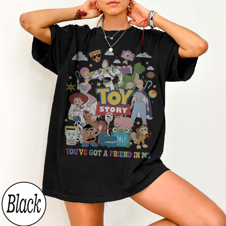 May include: A black short-sleeve t-shirt featuring a colorful graphic print of Toy Story characters including Buzz Lightyear, Woody, and Jessie. The design includes the text TOY STORY and YOU'VE GOT A FRIEND IN ME in multi-colored lettering.