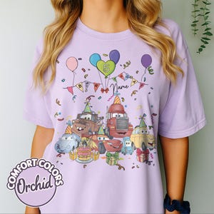 Peut inclure: T-shirt lilas Comfort Colors avec un motif d'anniversaire. Le motif pr&eacute;sente des voitures de dessins anim&eacute;s, des ballons, des confettis et un g&acirc;teau d'anniversaire, avec le chiffre 5 et un espace pour un nom. Le t-shirt a un col rond et des manches courtes.