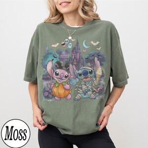 May include: A moss green short-sleeve t-shirt featuring a graphic print of two cartoon characters, one dressed as a pumpkin and the other as a mummy, set against a backdrop of a castle, bats, and a crescent moon under a night sky.