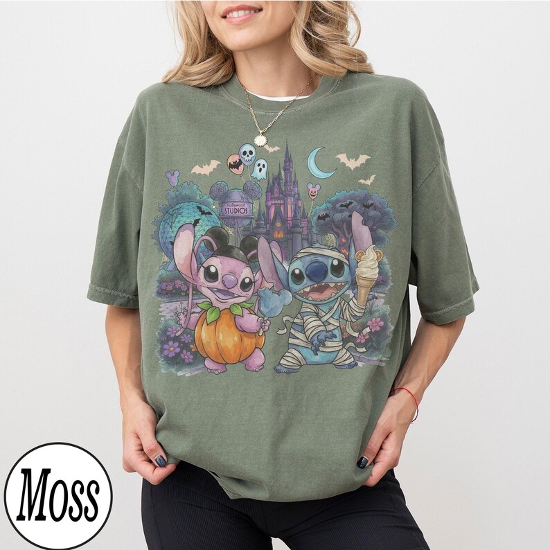 May include: A moss green short-sleeve t-shirt featuring a graphic print of two cartoon characters, one dressed as a pumpkin and the other as a mummy, set against a backdrop of a castle, bats, and a crescent moon under a night sky.