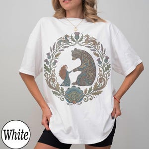 以下が含まれることがあります： 白のTシャツ。円形のデザインで、クマと女の子が手をつないでおり、花の装飾が施されています。デザインは茶色、青、緑の色合いを使用しています。「White」の文字が白い円で下部にあります。