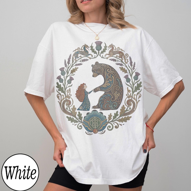 以下が含まれることがあります： 白のTシャツ。円形のデザインで、クマと女の子が手をつないでおり、花の装飾が施されています。デザインは茶色、青、緑の色合いを使用しています。「White」の文字が白い円で下部にあります。