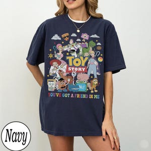 May include: A navy blue short-sleeve t-shirt featuring a colorful graphic print of Toy Story characters including Buzz Lightyear, Woody, and Jessie. The text Toy Story and You've Got A Friend In Me are printed in the center with various icons.