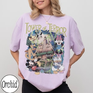K&ouml;nnte beinhalten: Helllila T-Shirt mit einem Aufdruck des Tower of Terror und den Disney-Figuren Mickey Mouse, Minnie Mouse, Goofy und Donald Duck. Der Text "Tower of Terror" steht &uuml;ber dem Aufdruck. Das T-Shirt hat kurze &Auml;rmel.