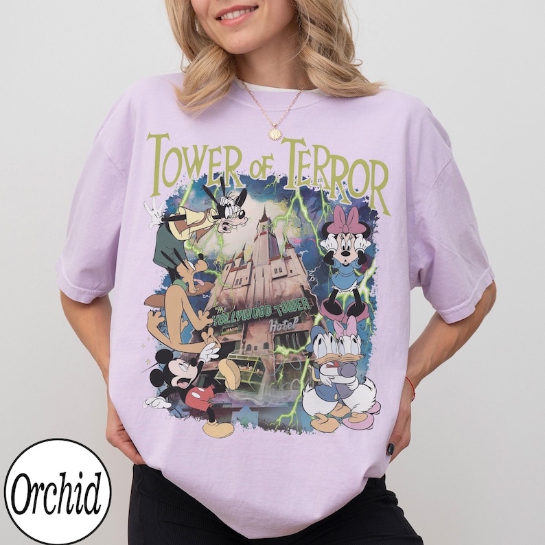 K&ouml;nnte beinhalten: Helllila T-Shirt mit einem Aufdruck des Tower of Terror und den Disney-Figuren Mickey Mouse, Minnie Mouse, Goofy und Donald Duck. Der Text "Tower of Terror" steht &uuml;ber dem Aufdruck. Das T-Shirt hat kurze &Auml;rmel.