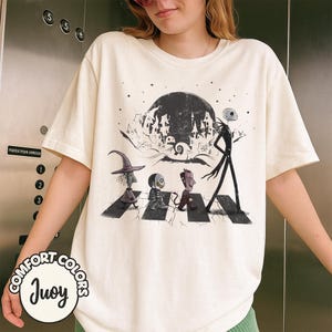 Peut inclure: T-shirt cr&egrave;me avec un motif de personnages de L'&Eacute;trange No&euml;l de Monsieur Jack traversant une rue, avec une pleine lune et une ville effrayante en arri&egrave;re-plan. Le t-shirt porte les mots "Comfort Colors" et "Juoy".