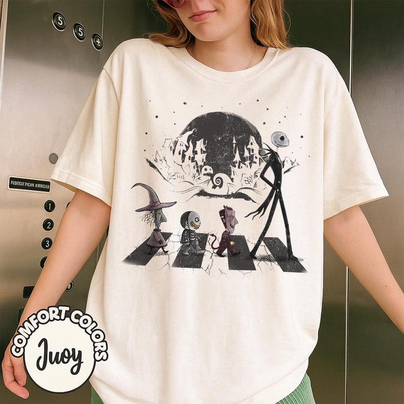 Peut inclure: T-shirt cr&egrave;me avec un motif de personnages de L'&Eacute;trange No&euml;l de Monsieur Jack traversant une rue, avec une pleine lune et une ville effrayante en arri&egrave;re-plan. Le t-shirt porte les mots "Comfort Colors" et "Juoy".