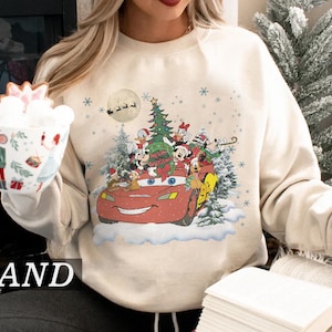 K&ouml;nnte beinhalten: Sandfarbenes Sweatshirt mit festlichem Design. Disney-Figuren wie Mickey Mouse und Donald Duck fahren in einem roten Lightning McQueen-Auto. Ein Weihnachtsbaum und Schneeflocken erg&auml;nzen die festliche Szene.