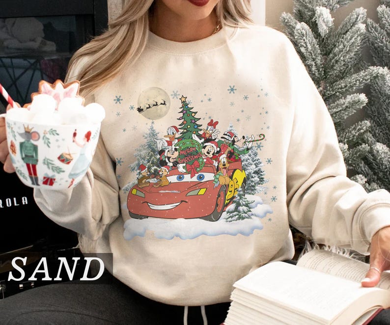 K&ouml;nnte beinhalten: Sandfarbenes Sweatshirt mit festlichem Design. Disney-Figuren wie Mickey Mouse und Donald Duck fahren in einem roten Lightning McQueen-Auto. Ein Weihnachtsbaum und Schneeflocken erg&auml;nzen die festliche Szene.