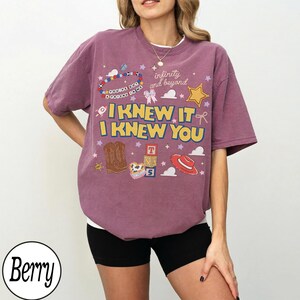 May include: A berry-colored short-sleeve t-shirt featuring a graphic print with the text "I KNEW IT I KNEW YOU" and "infinity and beyond". The design includes illustrations of cowboy boots, a red hat, alphabet blocks, and a beaded bracelet.