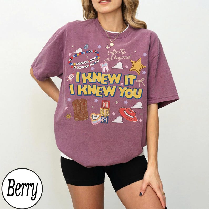 May include: A berry-colored short-sleeve t-shirt featuring a graphic print with the text "I KNEW IT I KNEW YOU" and "infinity and beyond". The design includes illustrations of cowboy boots, a red hat, alphabet blocks, and a beaded bracelet.