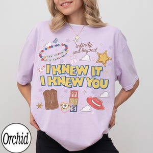 May include: A light purple t-shirt featuring a graphic print with the text "I KNEW IT I KNEW YOU", "infinity and beyond", and "YOU'VE GOT A FRIEND IN ME". The design includes illustrations of a beaded bracelet, cowboy boots, blocks, and a hat.