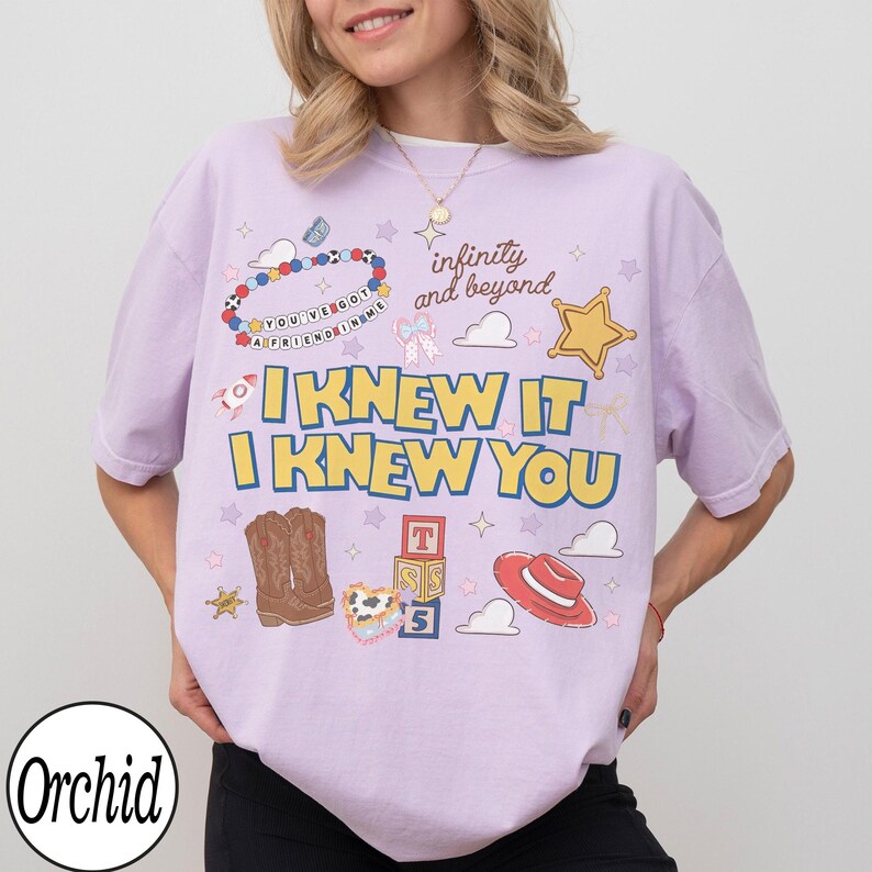 May include: A light purple t-shirt featuring a graphic print with the text "I KNEW IT I KNEW YOU", "infinity and beyond", and "YOU'VE GOT A FRIEND IN ME". The design includes illustrations of a beaded bracelet, cowboy boots, blocks, and a hat.