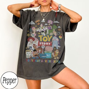 May include: A charcoal gray short-sleeve t-shirt featuring a colorful graphic print of Toy Story characters including Buzz Lightyear, Woody, and Jessie. The design includes the text TOY STORY and YOU'VE GOT A FRIEND IN ME in a retro font style.
