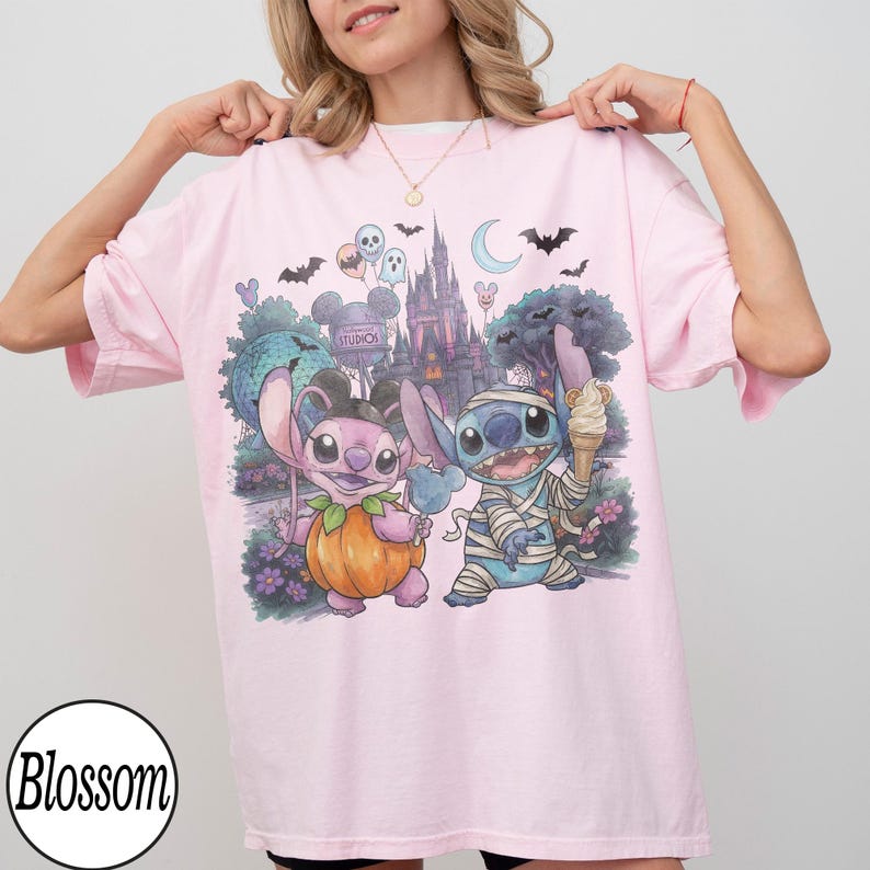 May include: A light pink t-shirt featuring a graphic print of two blue and pink alien characters in Halloween costumes. One character is dressed as a pumpkin and the other as a mummy, standing in front of a castle, bats, and balloons.