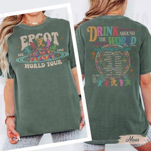 May include: Green t-shirt with "EPCOT World Tour" graphic on the front, featuring a retro design with the year 1982. The back of the shirt says "Drink Around the World" with a globe graphic and country names. The shirt is a moss green color.