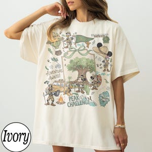 May include: Ivory t-shirt with a colourful graphic featuring cartoon characters and a safari theme. The design includes a hot air balloon, a jeep, and a tree, with the text "Adventure Awaits!" and "Peak Challenge".