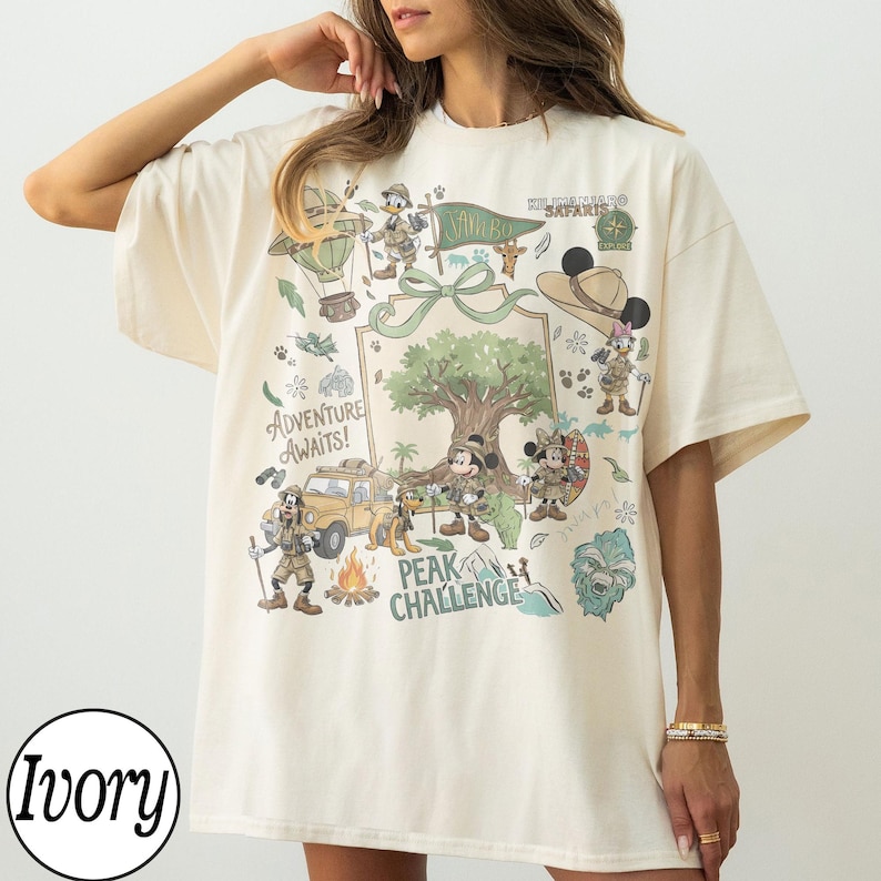 May include: Ivory t-shirt with a colourful graphic featuring cartoon characters and a safari theme. The design includes a hot air balloon, a jeep, and a tree, with the text "Adventure Awaits!" and "Peak Challenge".