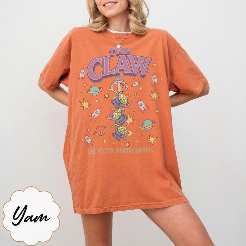 Puede incluir: Una camiseta naranja de manga corta con un estampado gr&aacute;fico que incluye el texto THE CLAW y THE MYSTIC PORTAL AWAITS. El dise&ntilde;o muestra tres alien&iacute;genas verdes de tres ojos colgados de una garra, rodeados de cohetes, planetas y estrellas.