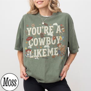 May include: A moss green short-sleeve t-shirt featuring the text "YOU'RE A COWBOY LIKE ME" in a cream-colored font. The design includes various illustrations of cowboy-themed items, stars, clouds, and characters from the Toy Story franchise.