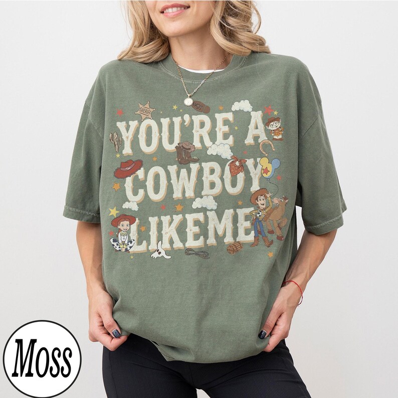 May include: A moss green short-sleeve t-shirt featuring the text "YOU'RE A COWBOY LIKE ME" in a cream-colored font. The design includes various illustrations of cowboy-themed items, stars, clouds, and characters from the Toy Story franchise.