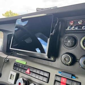 May include: Black tablet mounted on a dashboard, with a stylus resting on top. The dashboard features gauges, buttons, and switches. A charging cable is connected to the tablet.