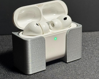 May include: White wireless earbuds in an open white charging case, held in a gray holder. The case has a green light illuminated. The earbuds are in the holder on a dark surface.