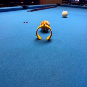 Puede incluir: Un soporte amarillo para bolas de billar rodea una bola de billar a rayas sobre una mesa de billar azul. Una bola blanca descansa en el fondo, con un taco de billar visible. La escena está ambientada en una habitación con poca luz.