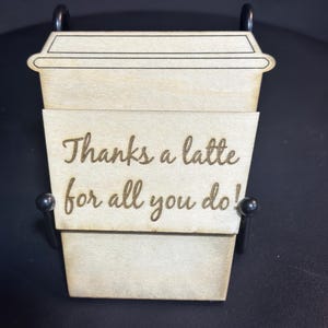 Könnte beinhalten: Eine Kaffeetasse aus Holz mit einem schwarzen Metallständer. Die Tasse trägt die Gravur "Thanks a latte for all you do!"