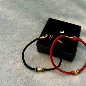 Puede incluir: Dos pulseras, una negra y otra roja, con detalles dorados y cierre de gancho. Las pulseras se muestran en una caja negra.