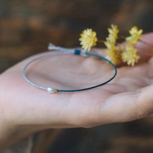 Könnte beinhalten: Ein zartes Armband aus blauem und grauem Garn mit einem kleinen Perlenanhänger.