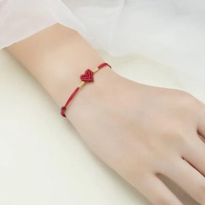 Può includere: Un braccialetto di corda rosso con un piccolo ciondolo a forma di cuore rosso. Il braccialetto è legato al polso di una persona.