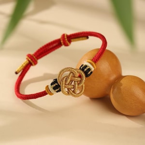 Puede incluir: Una pulsera roja y dorada con un diseño de nudo tejido. La pulsera presenta un nudo dorado central, acentuado con detalles en blanco y negro. El cordón rojo está anudado a intervalos, creando una pieza decorativa y simbólica.