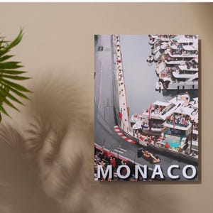 May include: A colorful aerial view of a race track in Monaco, with the word "MONACO" printed in white letters across the bottom of the image. The track is lined with white barriers and surrounded by a harbor filled with yachts and boats.