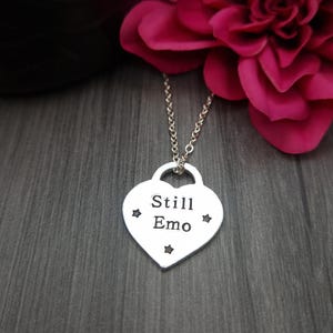 May include: Silver heart-shaped pendant necklace with a silver chain. The pendant has the words "Still Emo" engraved on it and four small stars around the text.