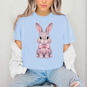 May include: Light blue t-shirt featuring a sparkly bunny graphic with a pink bow. The bunny has pink ears and is covered in glitter. The shirt is a soft, comfortable material.