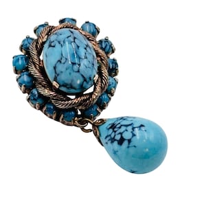 Puede incluir: Broche vintage con una gran piedra turquesa ovalada con vetas oscuras, rodeada por una cuerda de metal dorado retorcido y piedras turquesas más pequeñas. Una piedra turquesa en forma de lágrima cuelga debajo.