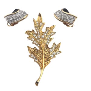 May include: A gold-toned leaf brooch and matching clip-on earrings set. The leaf brooch and earrings are encrusted with clear rhinestones. The earrings have a gold-toned frame with a black accent.