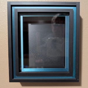 May include: A layered shadow box with a black frame and teal blue accents. The box has a glass front and a textured black interior. The frame is square and has a modern design, suitable for displaying art or collectibles.