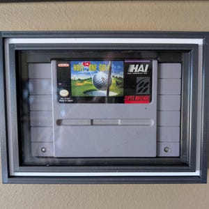 May include: A gray Super Nintendo video game cartridge titled Hole In One Golf by HAL America Inc. is displayed inside a black rectangular shadow box frame with a white inner border. The game label features a golf ball and a green golf course.