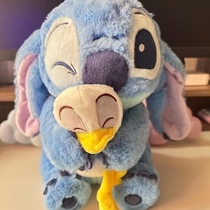 May include: Blue Stitch plush toy with a winking face, holding a yellow plush bird. The toy is made of soft, fuzzy material.