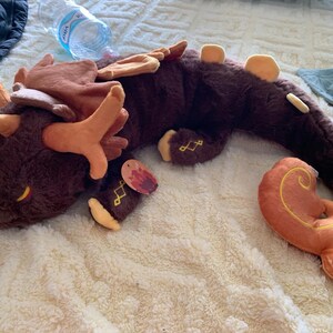 May include: A brown and orange plush dragon with a long tail and antlers. The dragon is lying on its side with its head resting on the ground. The dragon has a tag on its leg that says "Fire".