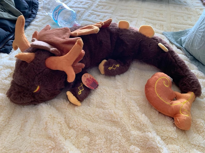 May include: A brown and orange plush dragon with a long tail and antlers. The dragon is lying on its side with its head resting on the ground. The dragon has a tag on its leg that says "Fire".