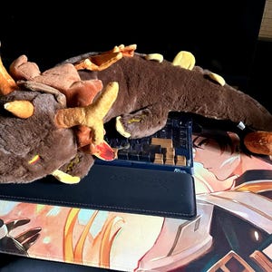 May include: A brown plush dragon with yellow and orange accents. The dragon is lying on a black keyboard and a mouse pad with a colorful anime design.