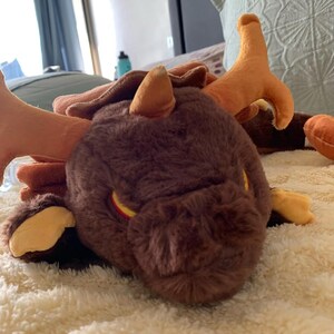 May include: A brown plush dragon with orange horns and yellow eyes. The dragon is lying on a white fluffy blanket.