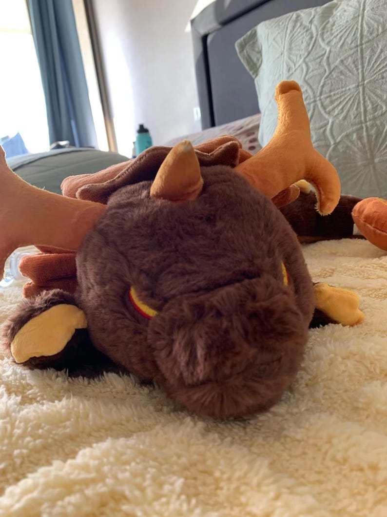 May include: A brown plush dragon with orange horns and yellow eyes. The dragon is lying on a white fluffy blanket.